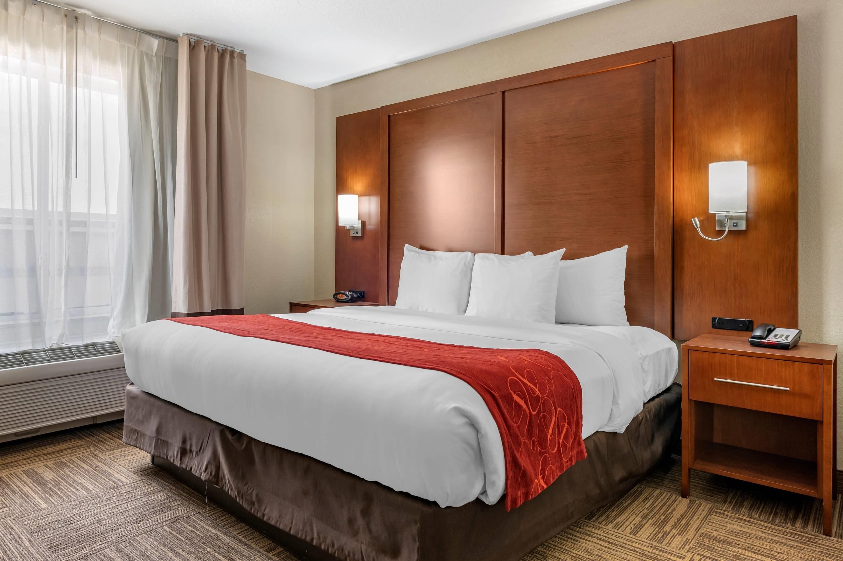 Quarto de hotel com uma cama grande, cabeceira de madeira, duas mesas de cabeceira com luminárias e uma janela com cortinas translúcidas.