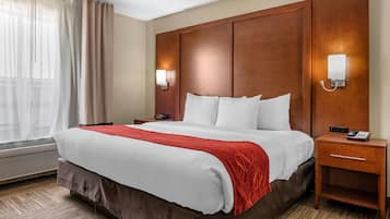 A hotel room with a large bed, a wooden headboard, two bedside tables with lamps, and a window with sheer curtains.