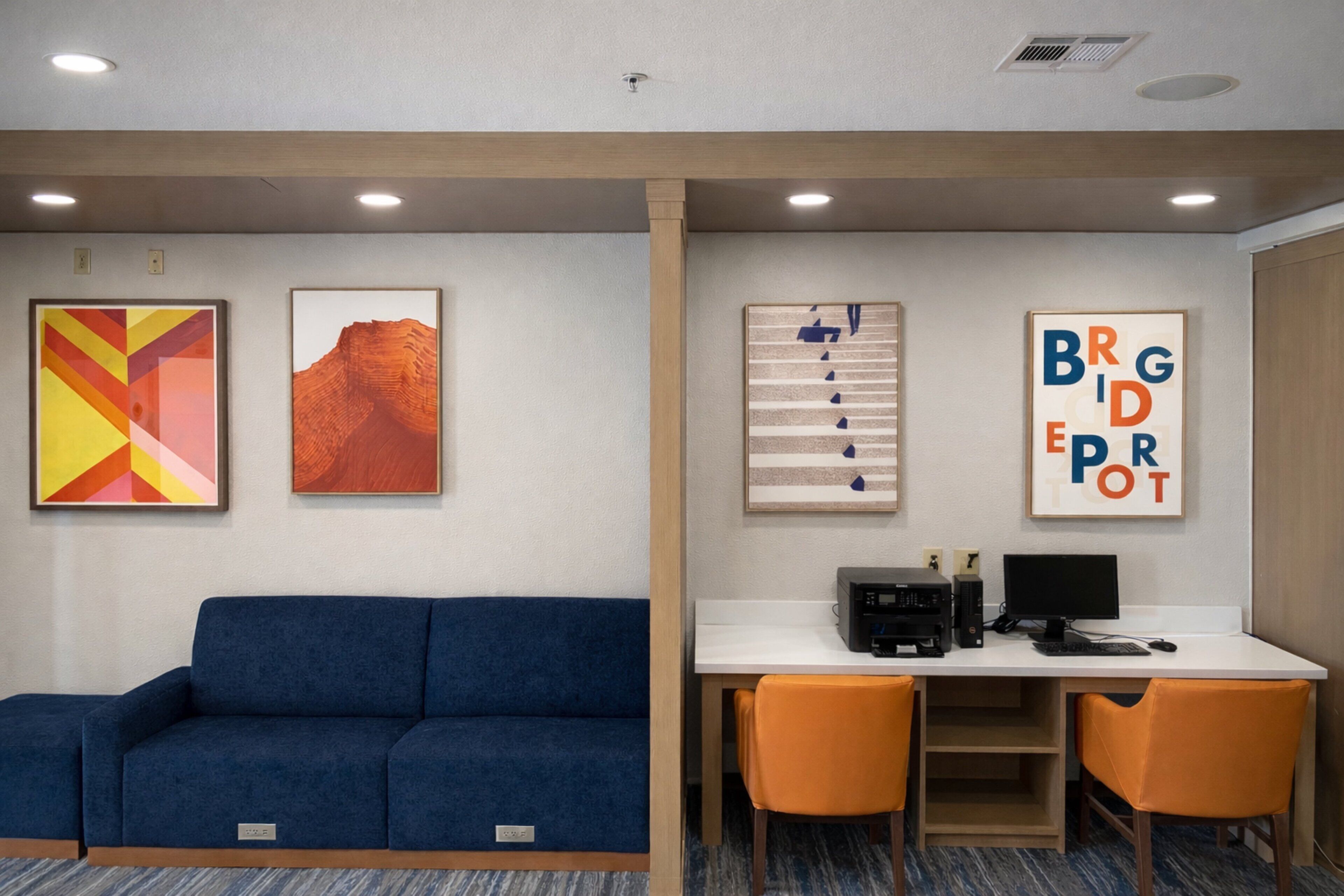 A room with a blue sofa, a desk with two orange chairs, and framed artwork on the walls.