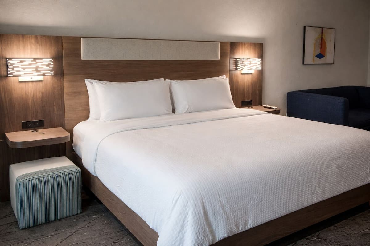 A modern hotel room with a large bed, wooden headboard, bedside tables, and a small stool.