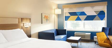 Quarto de hotel com uma cama grande, um sofá azul, uma poltrona amarela, uma mesa pequena e uma janela com cortinas estampadas em padrão geométrico.
