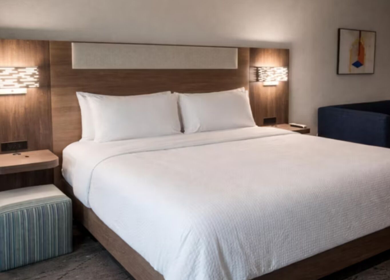 A hotel room with a large bed, two bedside tables, a nightstand, a chair, and wall-mounted lighting.