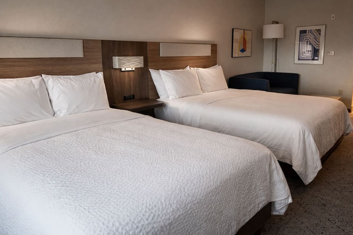 A hotel room with two beds, a bedside table, a lamp, and artwork on the wall.
