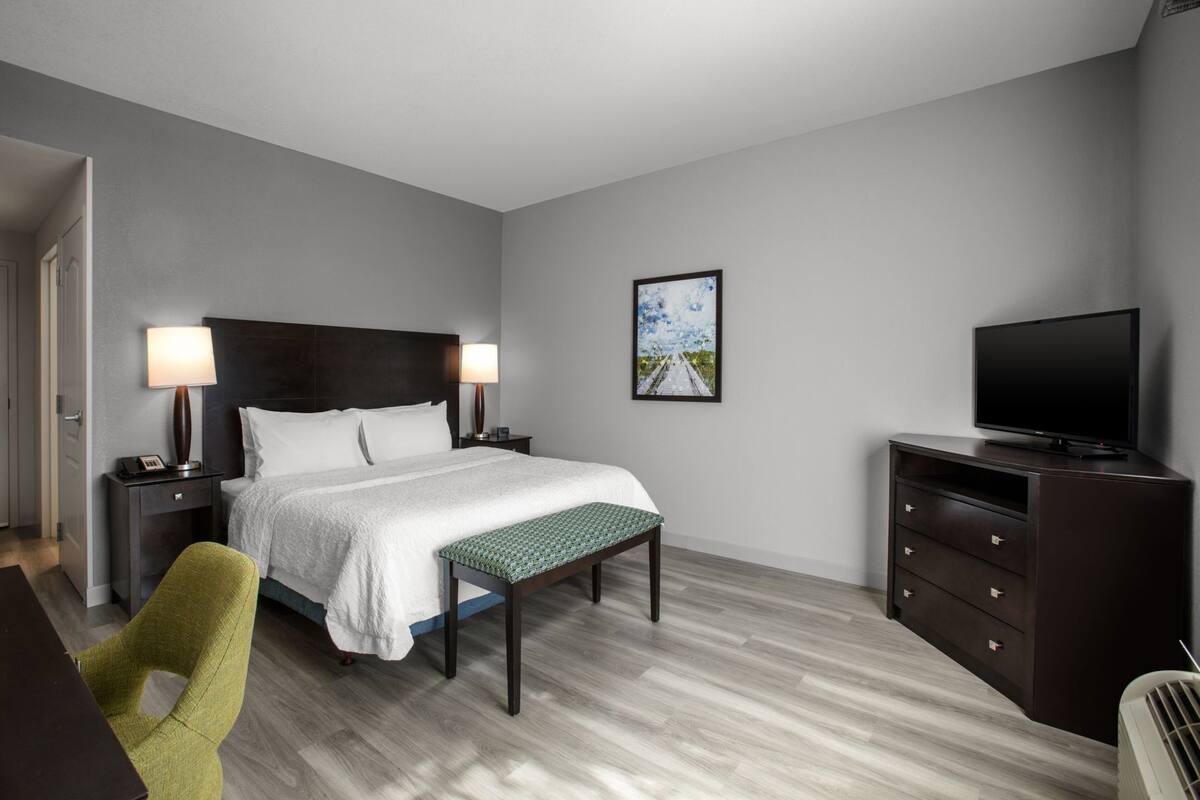 A hotel room with a bed, bedside tables with lamps, a television, a bench, a dresser, and a framed picture on the wall.