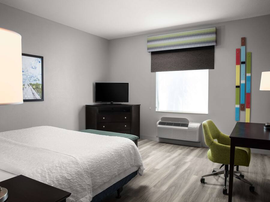 Habitación de hotel con cama, escritorio, silla, televisor y ventana con persianas.