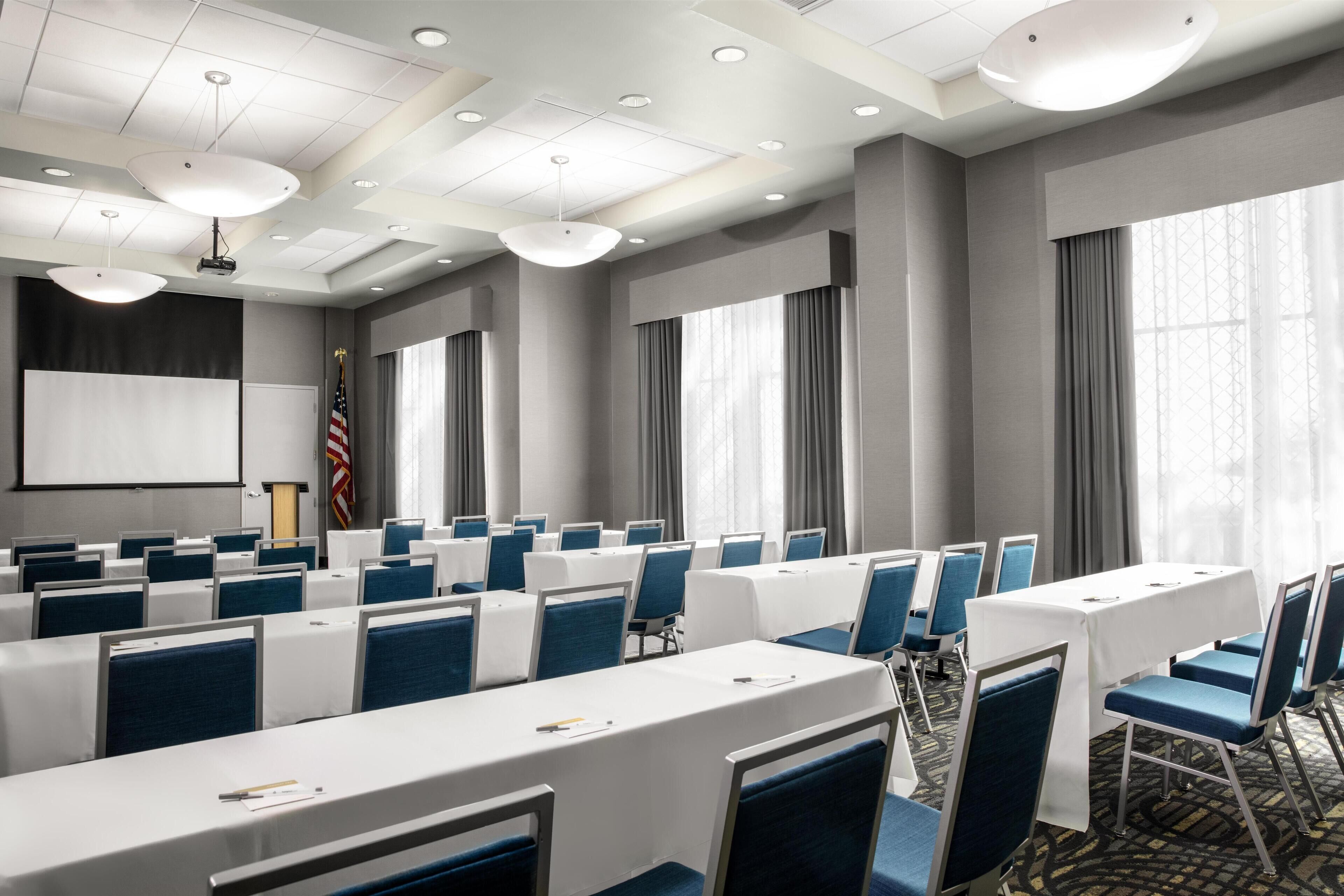 A conference room with rows of tables and chairs, a projector screen, and a podium with an American flag.