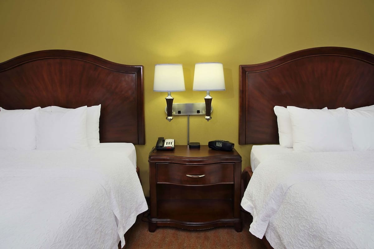 Quarto de hotel com duas camas de solteiro, mesa de cabeceira com abajur, telefone e relógio.