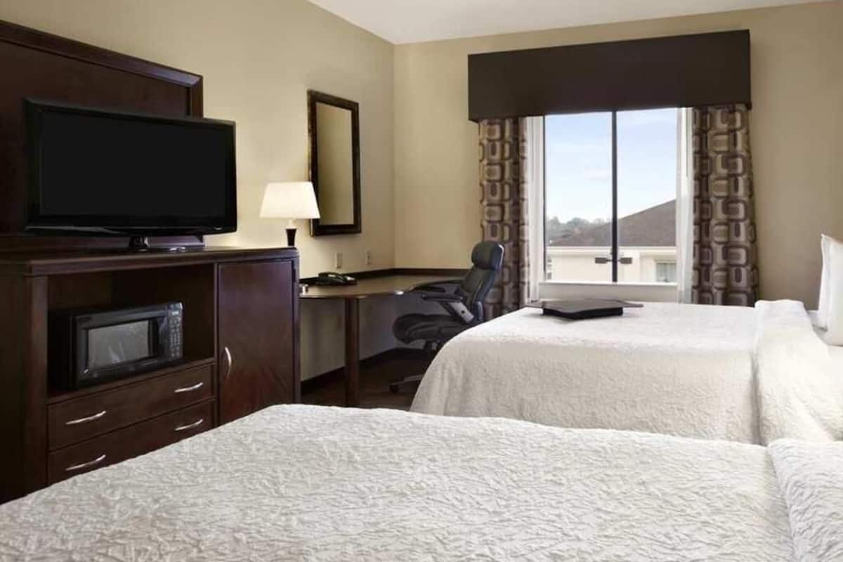 Quarto de hotel com duas camas, uma escrivaninha, uma cadeira, uma televisão e uma janela com cortinas.
