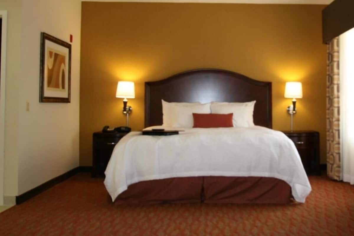 Quarto de hotel com uma cama grande, duas luminárias de cabeceira, uma mesa de apoio e um quadro na parede.