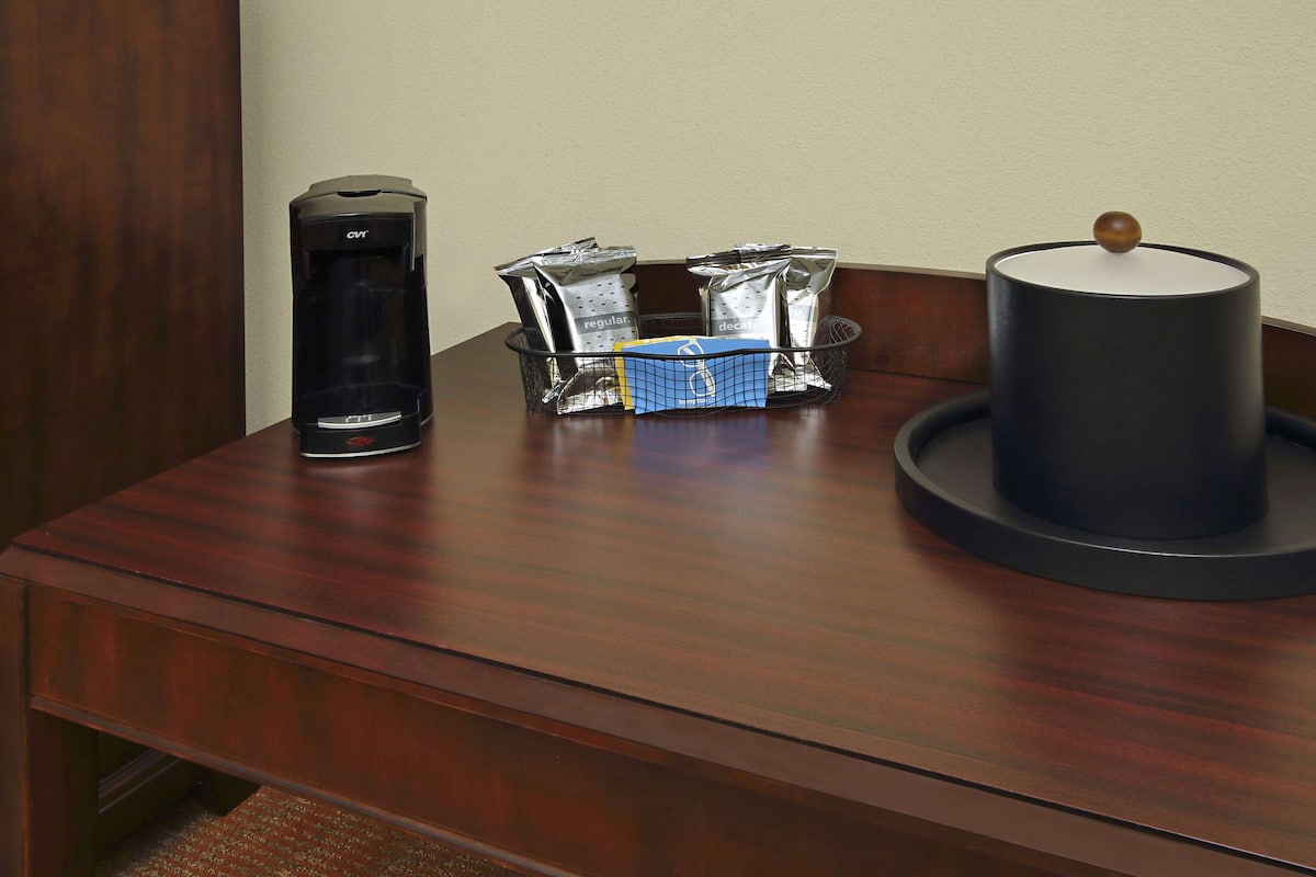 Uma mesa de madeira com uma cafeteira, uma cesta de embalagens de papel alumínio e um recipiente cilíndrico preto com um botão de madeira.