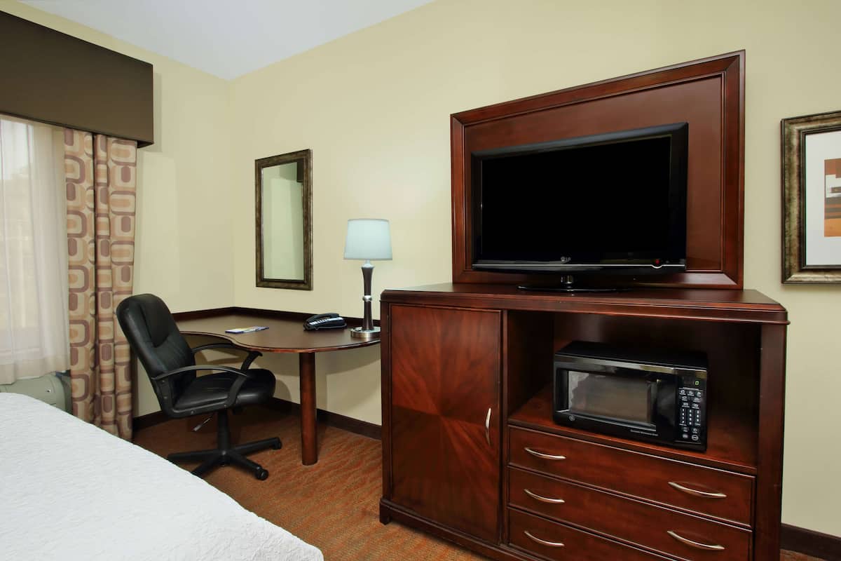 Quarto de hotel com cama, escrivaninha, cadeira, televisão em móvel de madeira e micro-ondas.