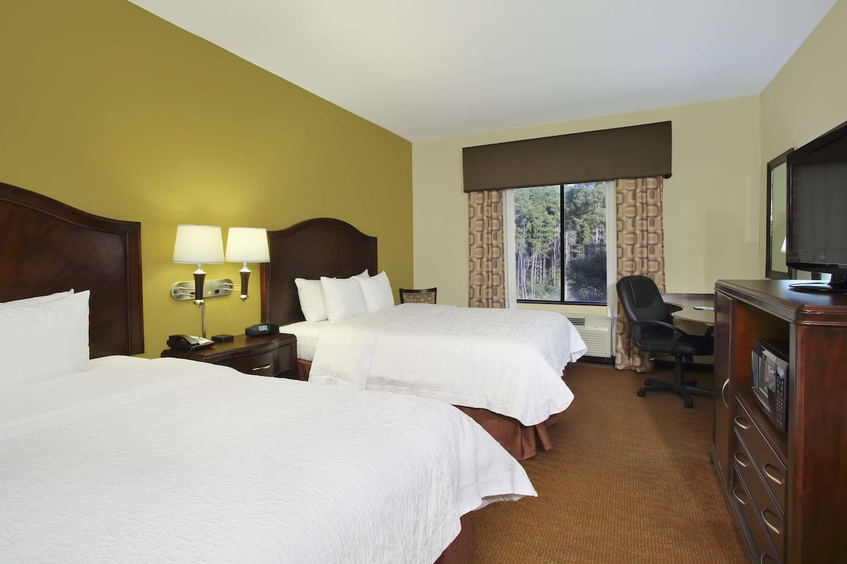Quarto de hotel com duas camas, uma escrivaninha, uma cadeira, televisão e janela com cortinas.