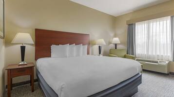 A hotel room with a large bed, two bedside tables with lamps, a chair, a window with curtains, and an air conditioning unit.