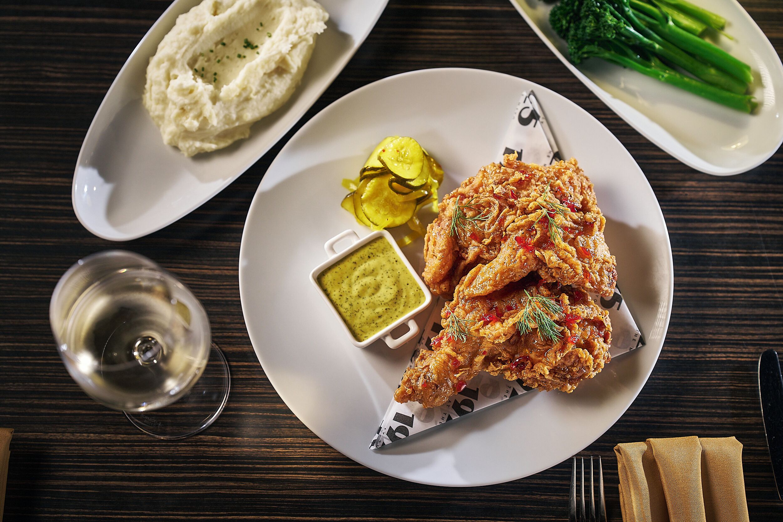 A plate of fried chicken with dipping sauce, mashed potatoes, pickles, and broccoli on a wooden table.