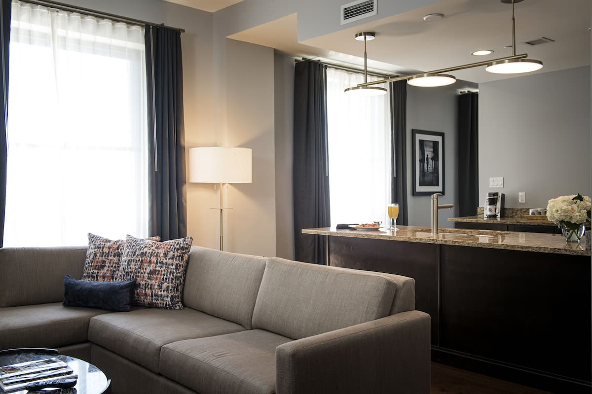 Ein modernes Hotelzimmer mit einer Couch, einem Couchtisch, einer Lampe, einer Küchenzeile mit Arbeitsfläche und einem großen Fenster mit Vorhängen.