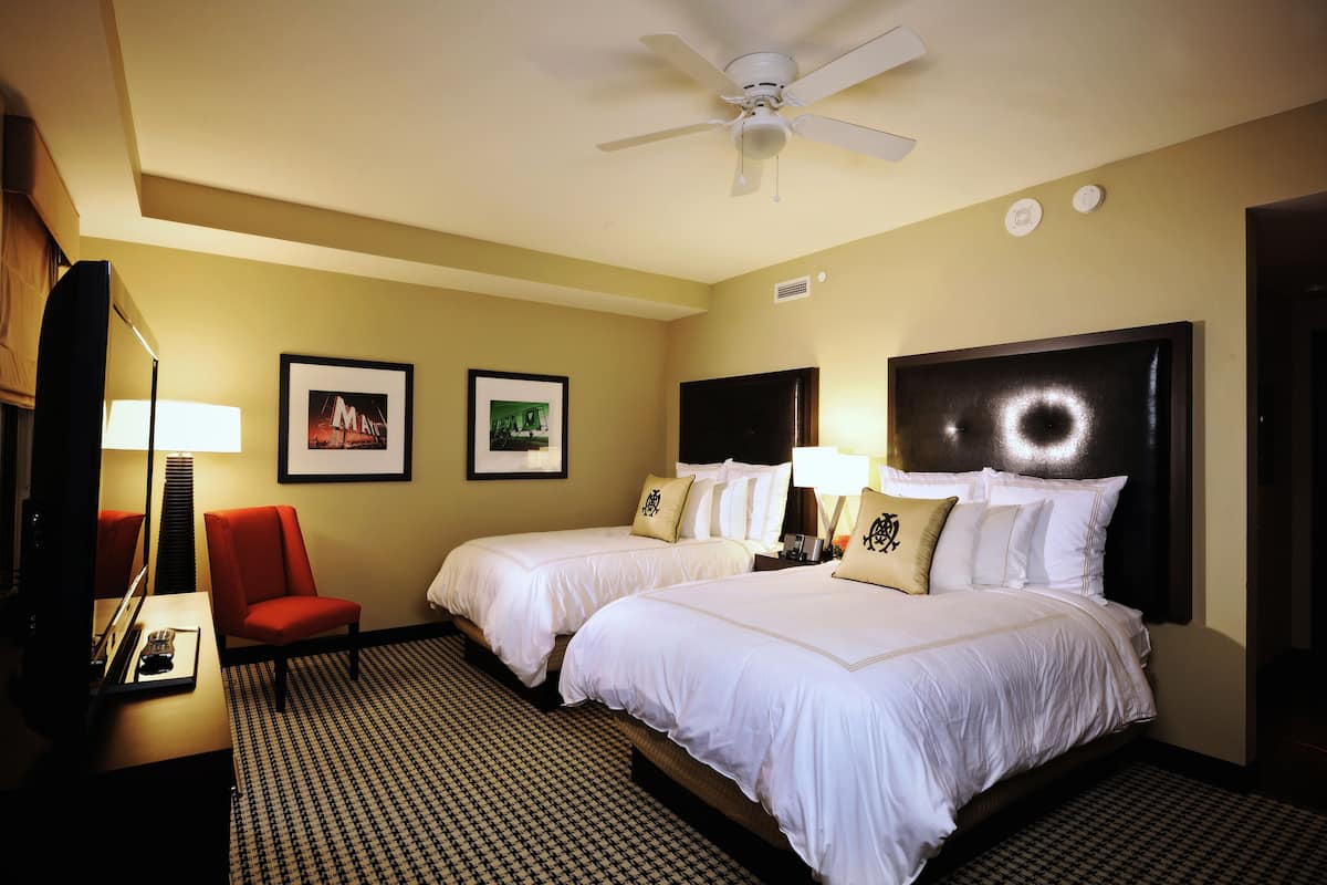 Ein Hotelzimmer mit zwei Betten, einem Deckenventilator, einem roten Sessel, einem Schreibtisch mit Lampe und gerahmten Bildern an der Wand.