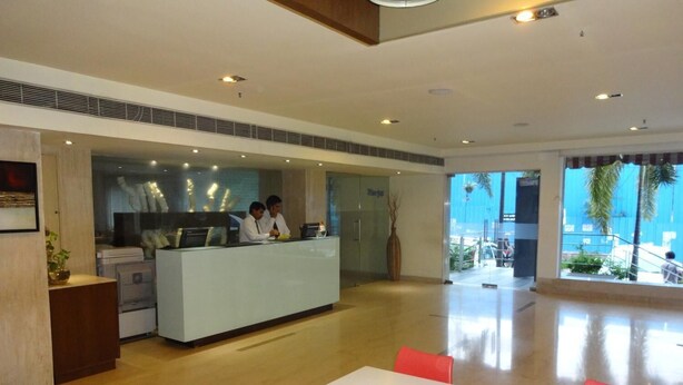 A modern hotel lobby with a reception desk, a dining area with a table and chairs, and large windows offering a view of the outdoors. - Minerva Grand Banjara Hills (Hyderabad)