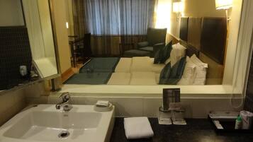 Un baño de hotel con un lavamanos grande, un espejo y vistas al área del dormitorio, que incluye una cama y una silla.