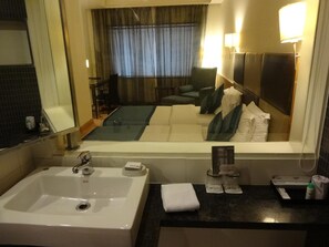 A hotel bathroom with a large sink, mirror, and a view of the bedroom area including a bed and a chair. - Minerva Grand Banjara Hills (Hyderabad)