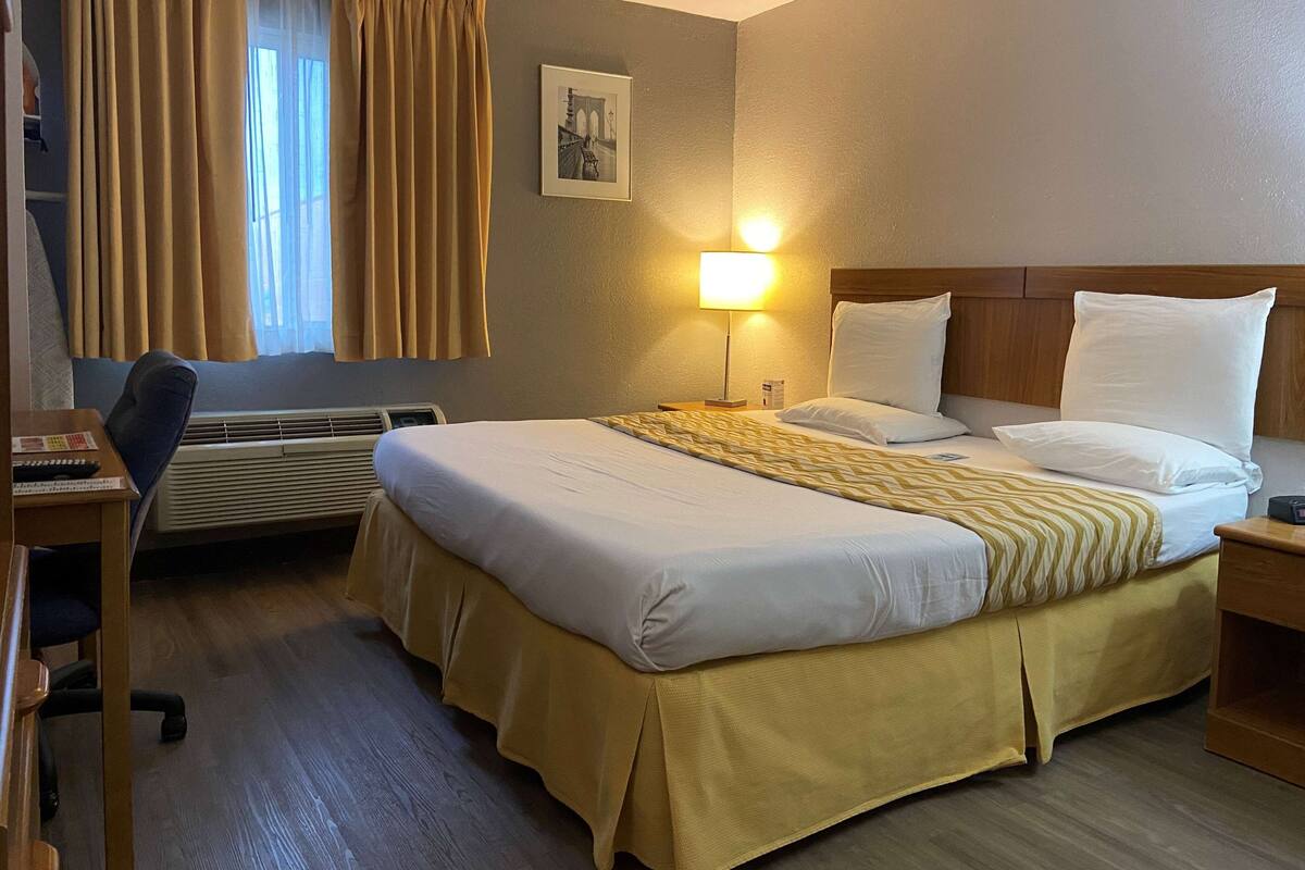 A hotel room with a bed, a desk, a chair, a lamp, and a window with curtains.
