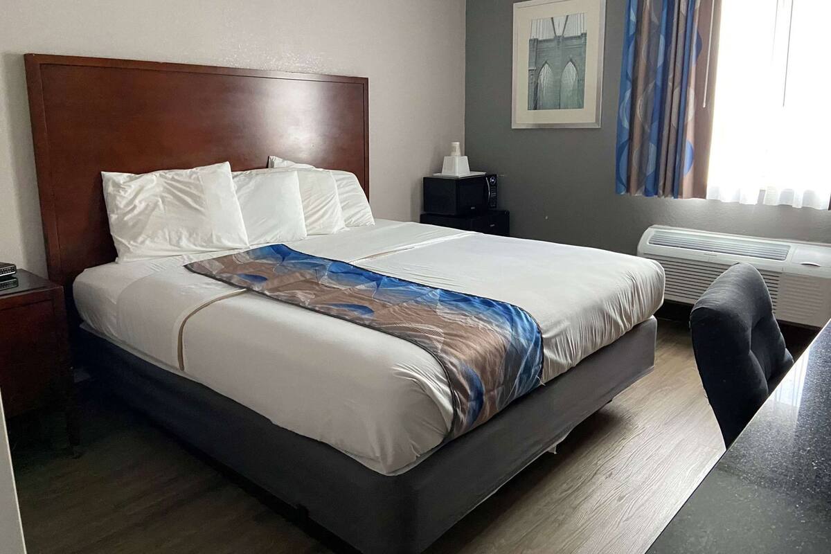 A hotel room with a large bed, a bedside table, a desk, a chair, a picture on the wall, and a window with curtains.