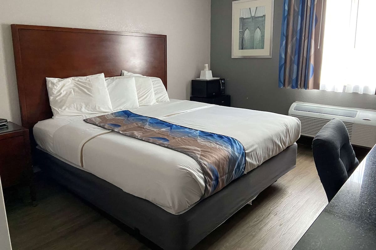 A hotel room with a large bed, a bedside table, a desk, a chair, a picture on the wall, and a window with curtains.