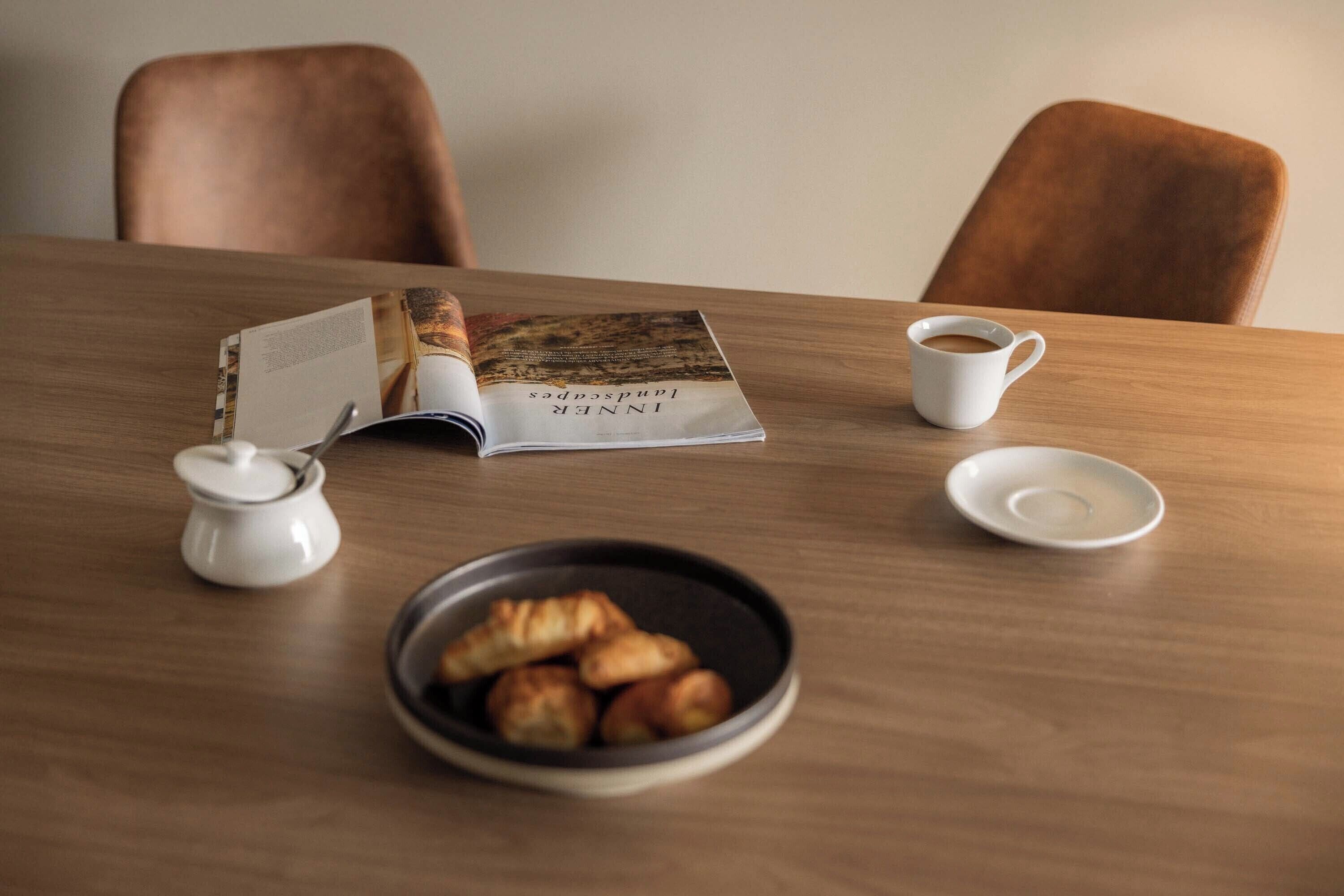 A wooden table with a magazine, a cup of coffee, a small bowl of croissants, and a sugar container.