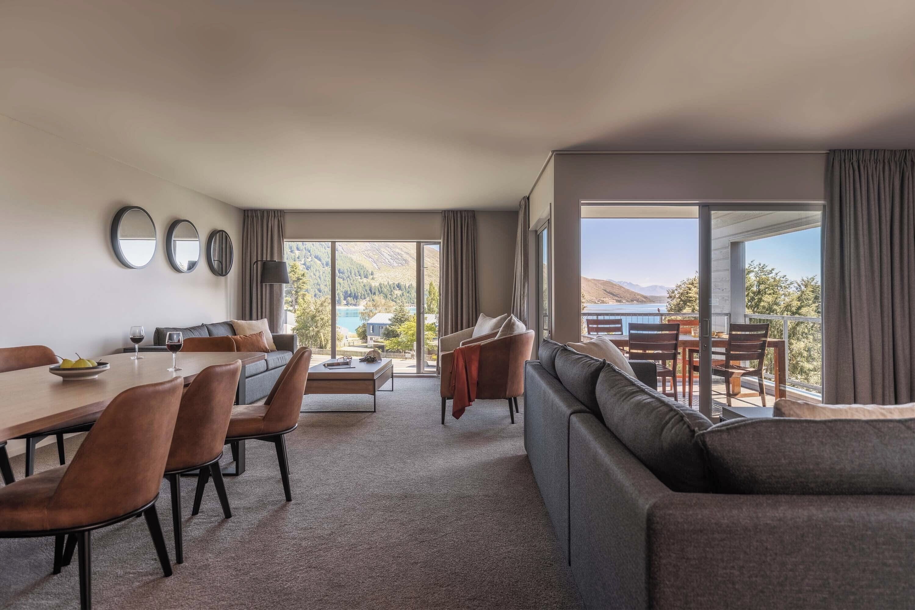 A modern hotel room with a dining area, a sofa, and a large window offering a view of the outdoors.