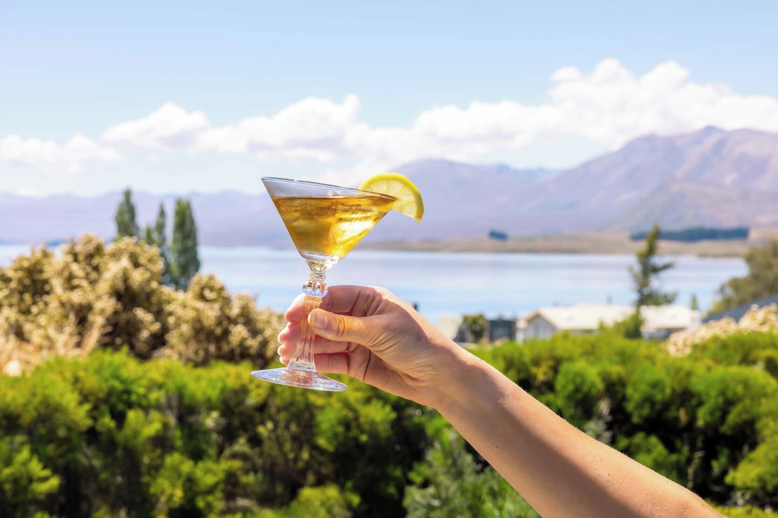 A hand holding a martini glass with a lemon slice, set against a scenic backdrop of mountains and a lake.