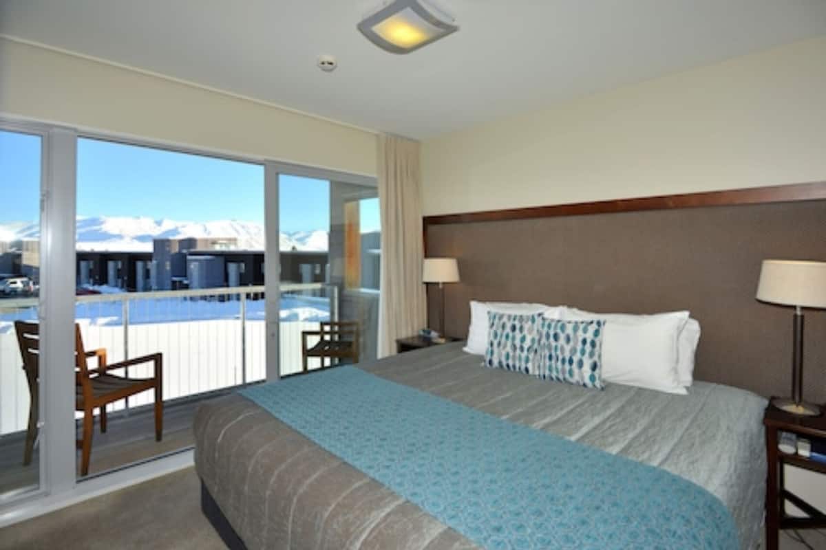 Habitación de hotel con una cama grande, dos mesitas de noche, un ventanal con vistas a montañas y un balcón con sillas.