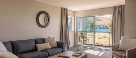 A modern living room with a sofa, armchair, and coffee table, a large window with a view of a mountainous landscape, and a circular mirror on the wall.