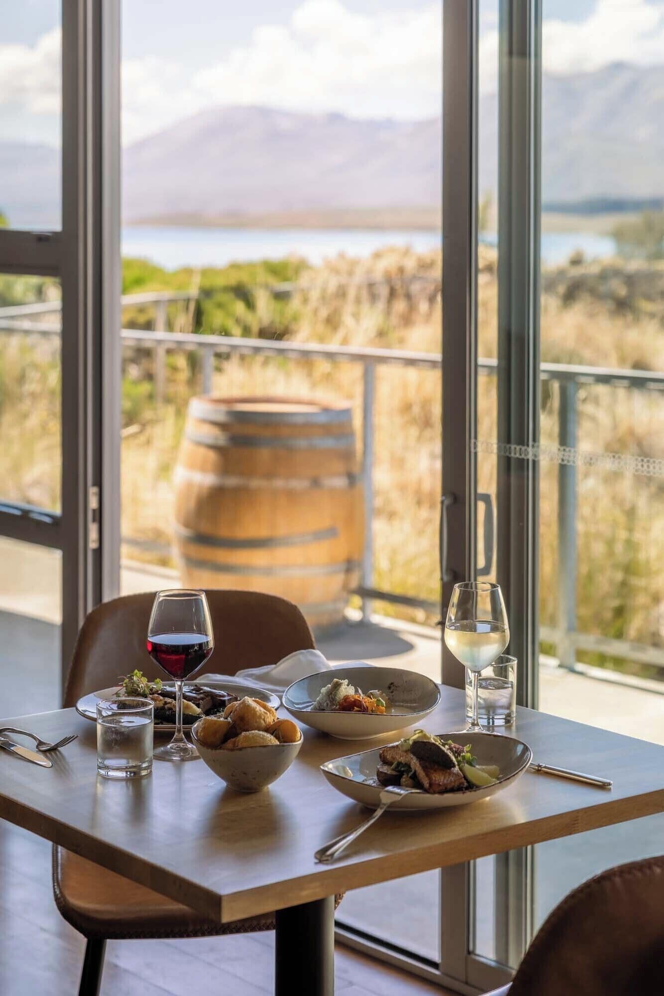 A dining table with plates of food, glasses of water and wine, and a view of a mountainous landscape through large windows.