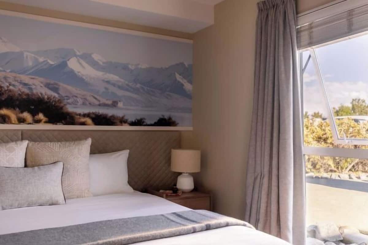 Un dormitorio con cama, mesita de noche con lámpara, ventana con cortinas y un mural de montañas en la pared.