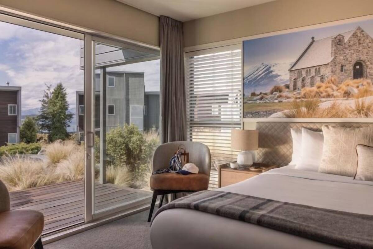 Habitación de hotel con cama, sillón, mesita de noche, lámpara, un cuadro y vistas a edificios y césped.