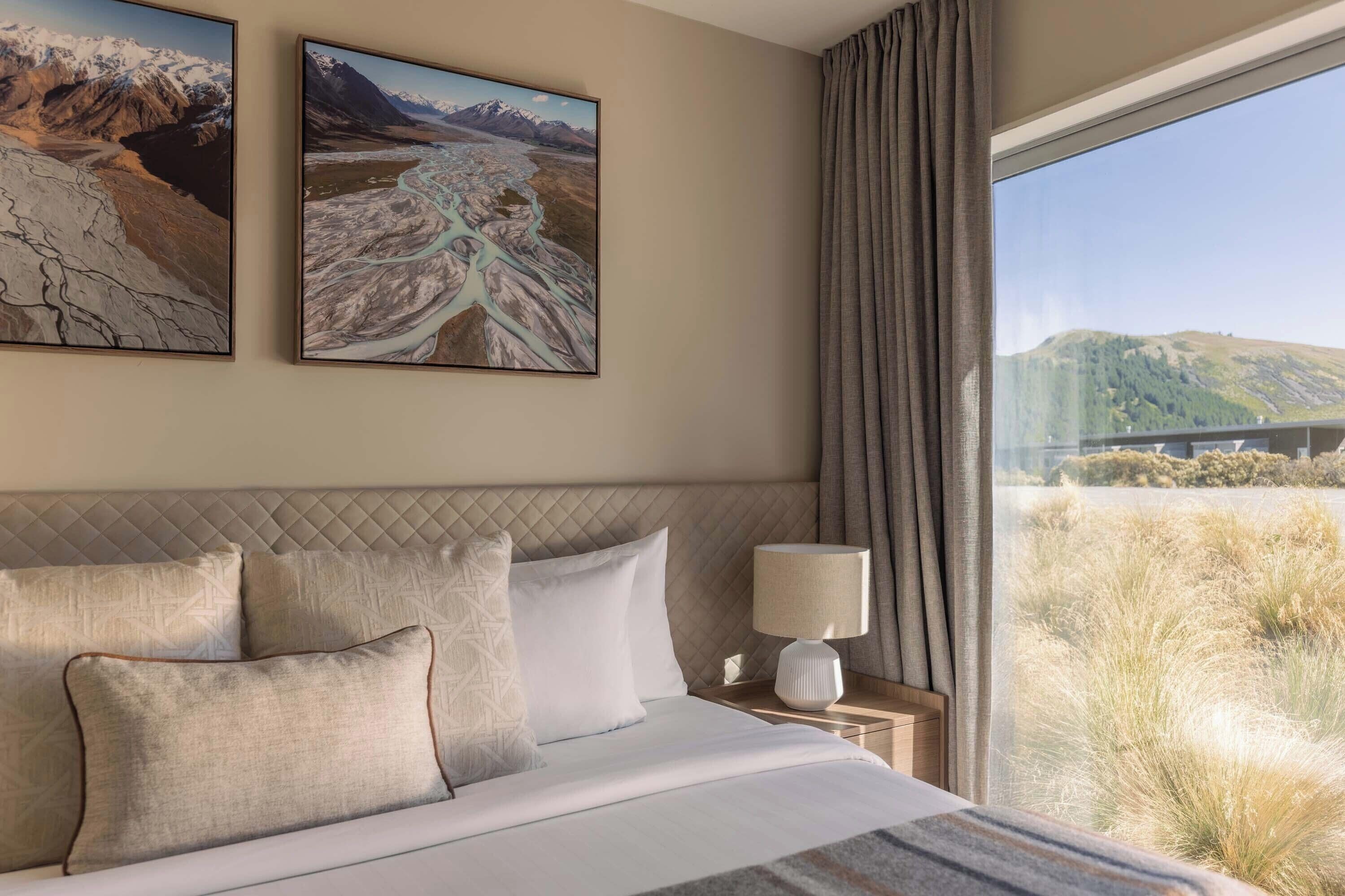 A hotel room with a large bed, a nightstand with a lamp, and two framed landscape paintings on the wall.