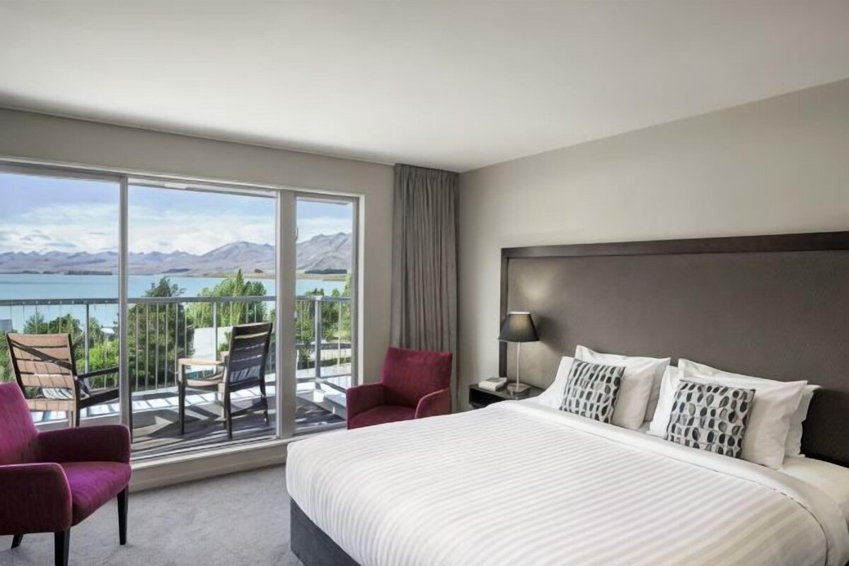 A hotel room with a large bed, a balcony with chairs, and a view of the mountains.