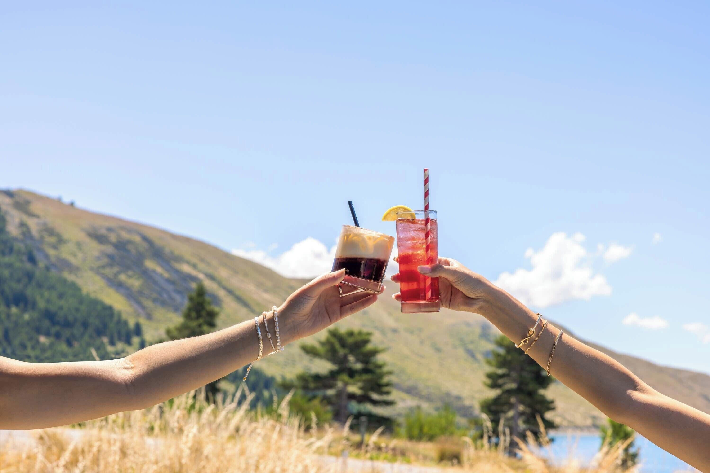 Dos manos sosteniendo vasos con bebidas con montañas al fondo.