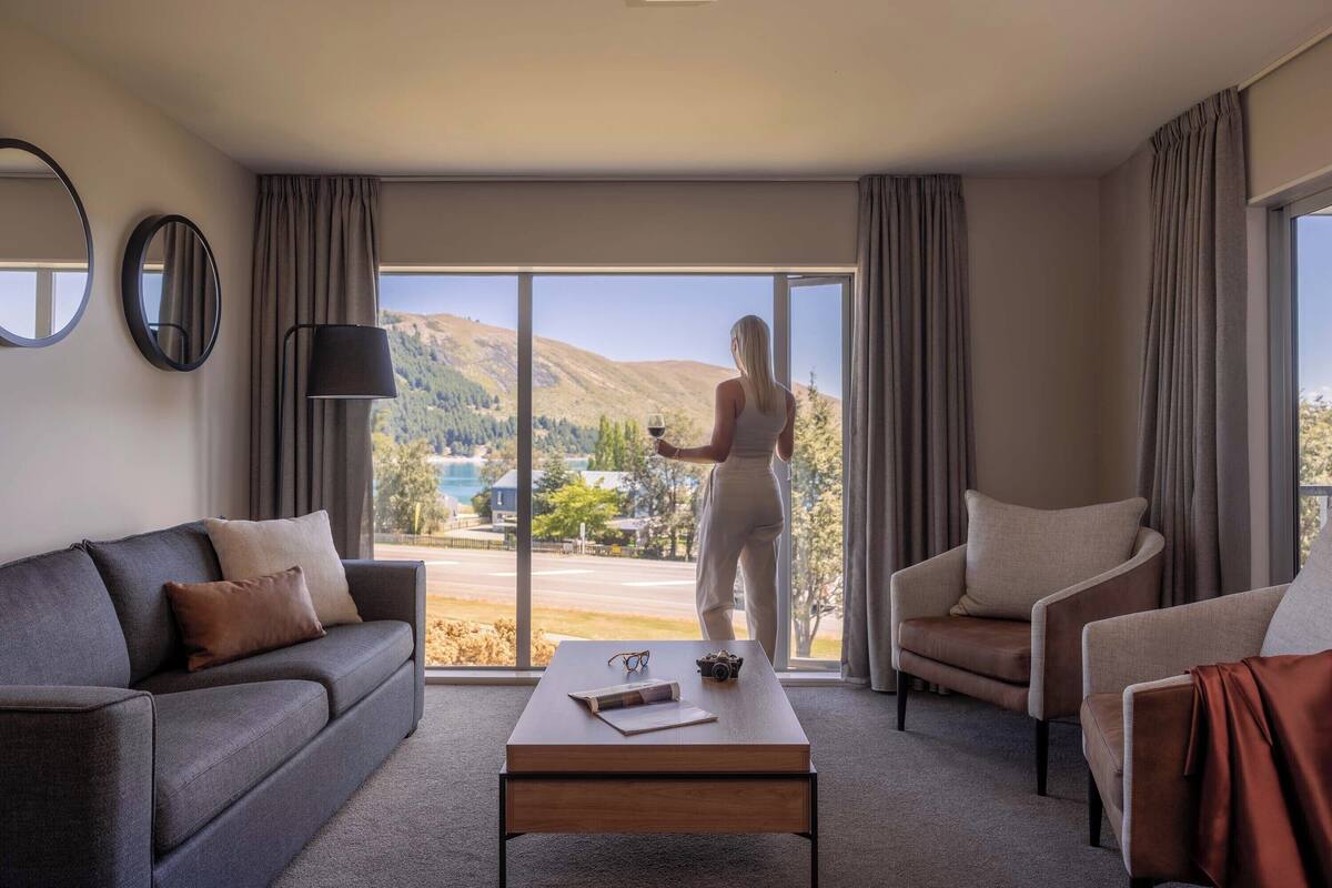 Habitación de hotel moderna con un amplio ventanal corredero que da a una vista panorámica, un sofá, una silla y una mesa de centro con un control remoto y vasos.