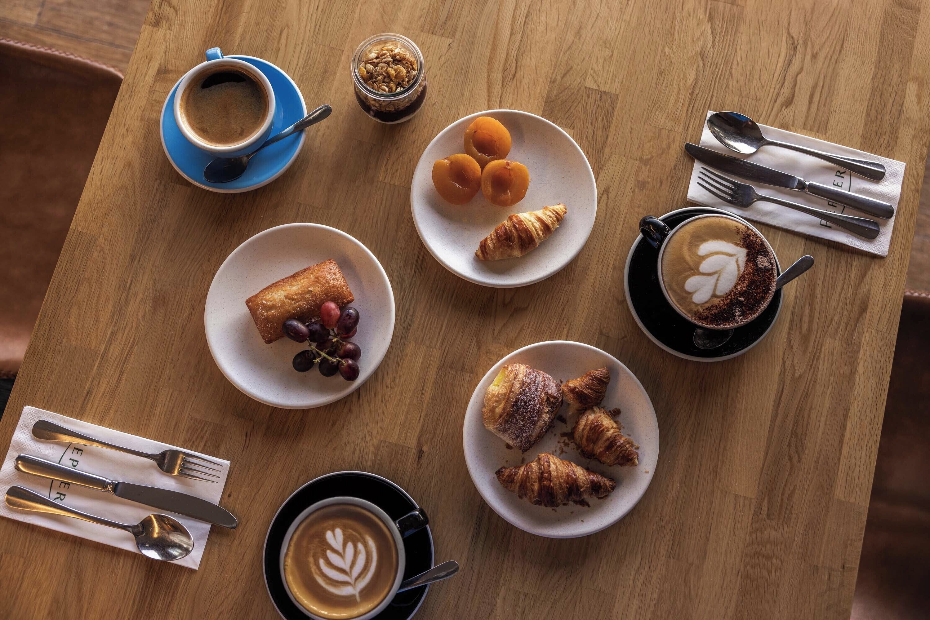 A wooden table with various breakfast items including croissants, a plate of grapes, and cups of coffee.