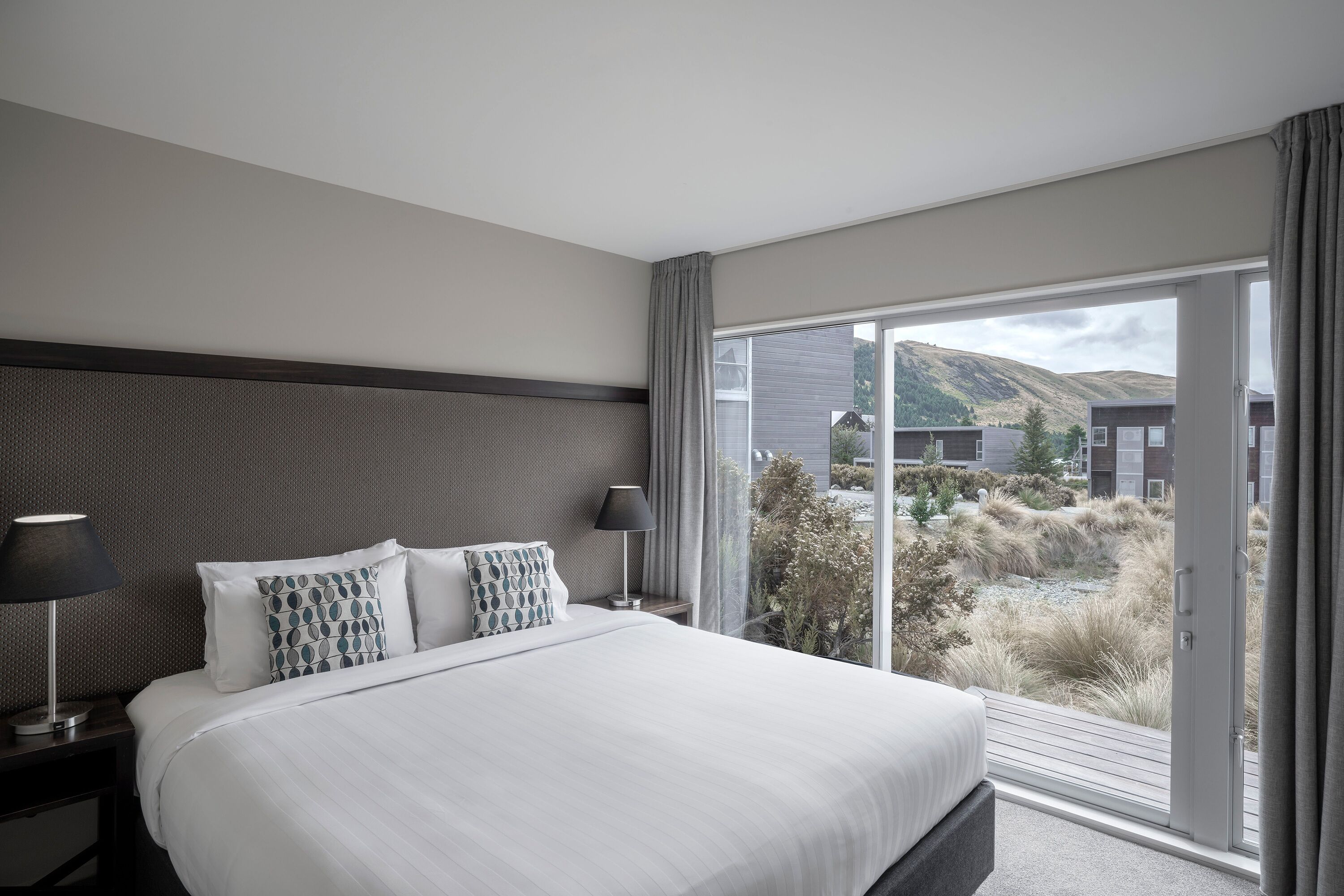 Et moderne hotelværelse med en stor seng, to sengeborde med lamper, samt en skydedør til en balkon med udsigt over bakkerne.