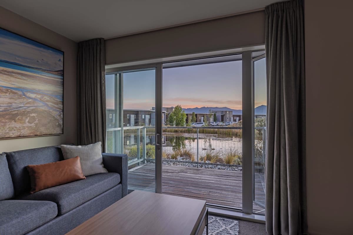 Una habitación de hotel moderna con un sofá gris, una mesa de centro de madera y un ventanal que ofrece vistas a un cuerpo de agua y montañas a lo lejos.