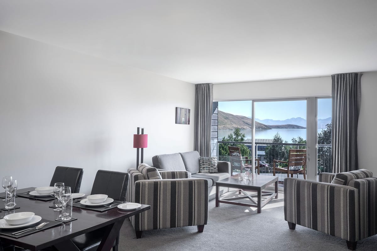 Una habitación de hotel moderna con zona de comedor, un conjunto de sofás y un ventanal con vistas a montañas y un lago.