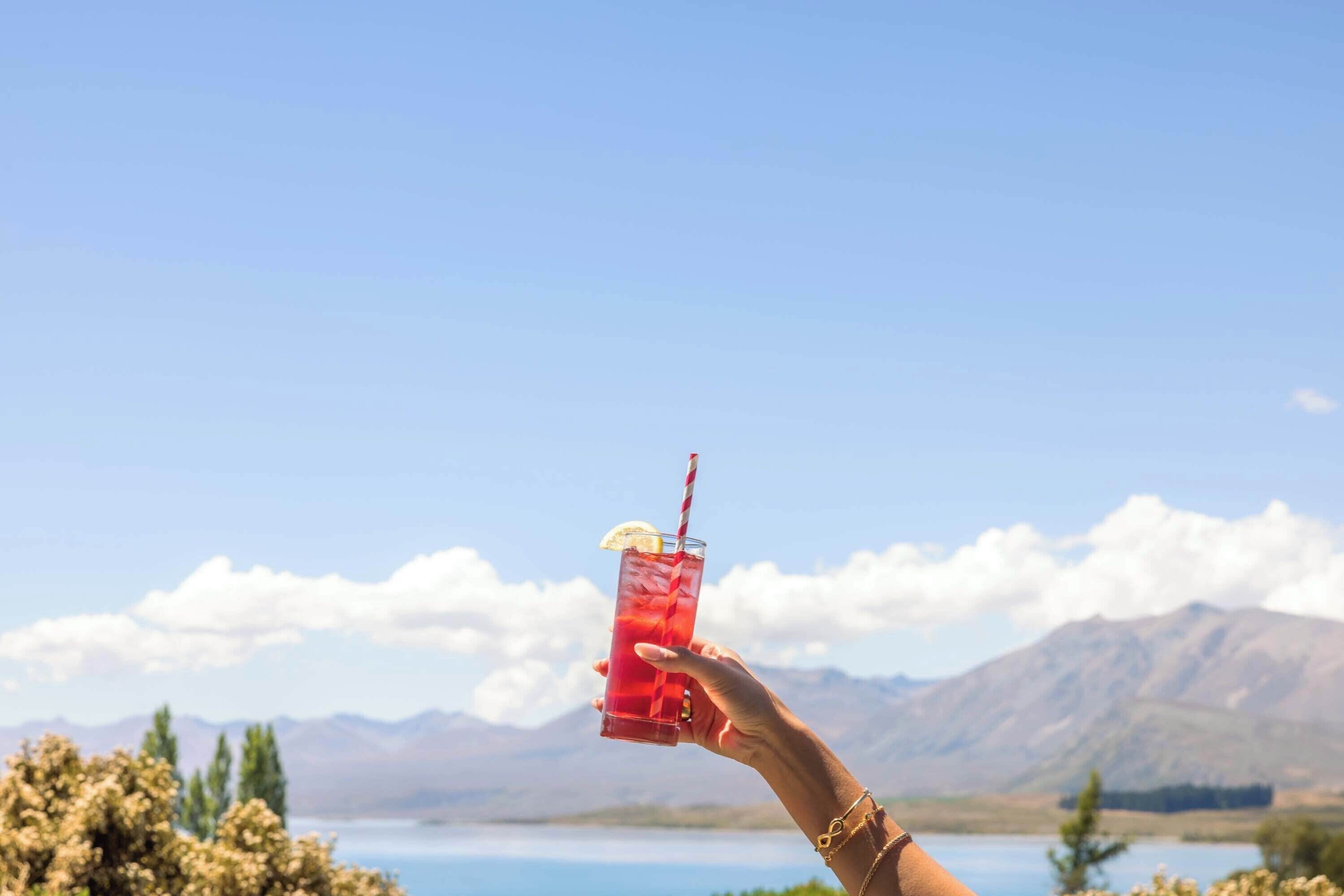 Une main tenant un verre de boisson rouge avec une paille, le tout sur fond d’un paysage montagneux sous un ciel dégagé.