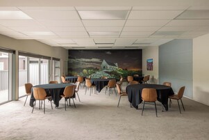 Un salón de conferencias con mesas y sillas, un gran mural en la pared y ventanas amplias.