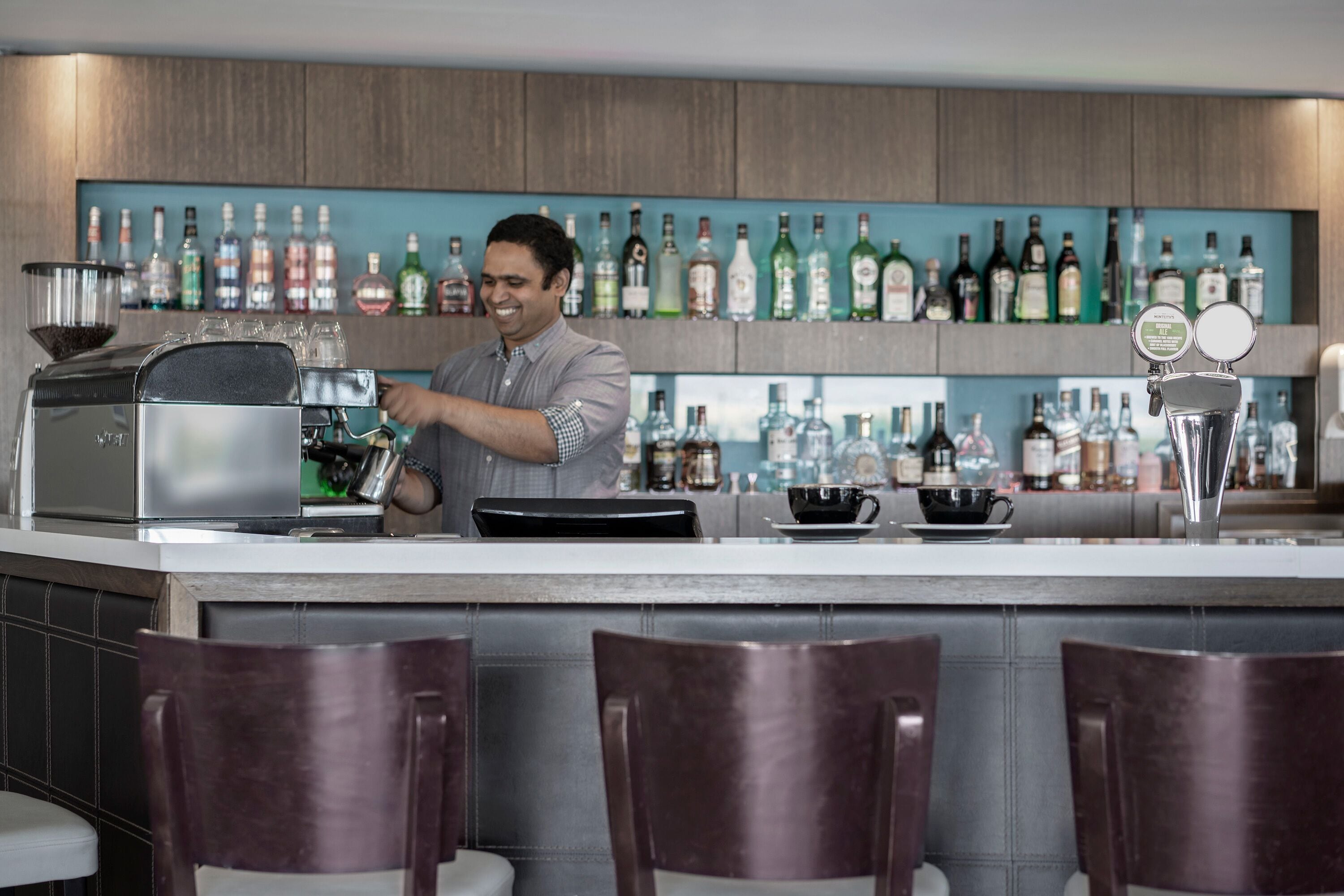 Zona de bar con una máquina de café, una estantería con diferentes botellas de licor y taburetes de bar.