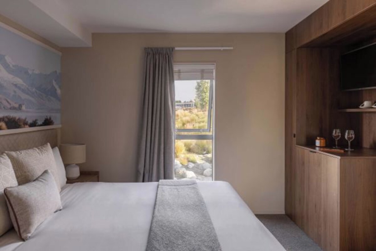 A hotel room with a bed, a window with curtains, a painting on the wall, a wooden cabinet, and a television.