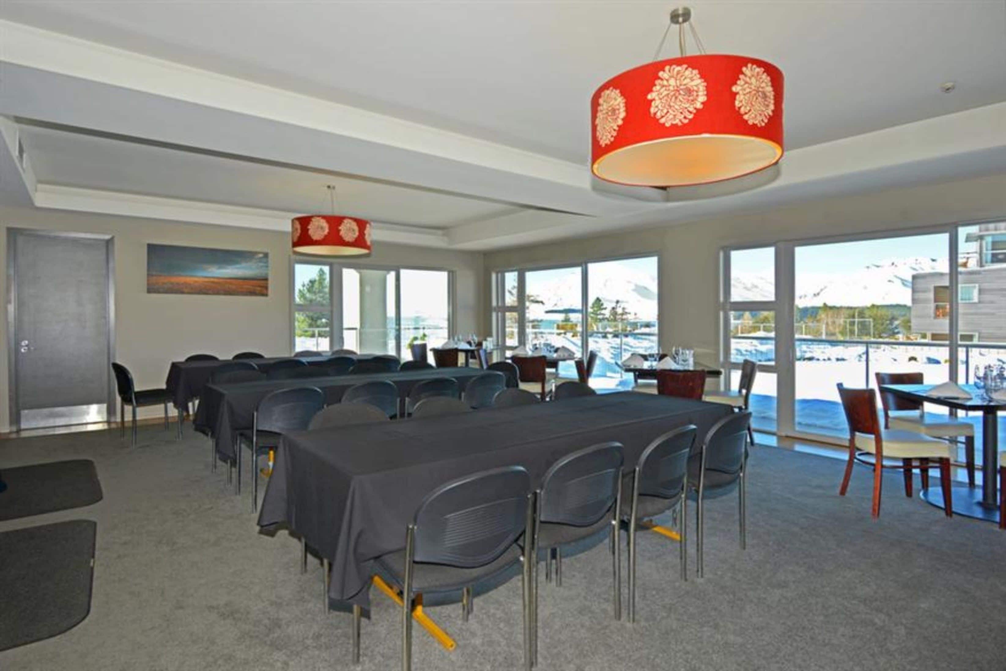 Une salle de conférence équipée de tables et de chaises, de grandes fenêtres offrant une vue sur un plan d’eau et des montagnes, ainsi que d’un luminaire décoratif rouge.