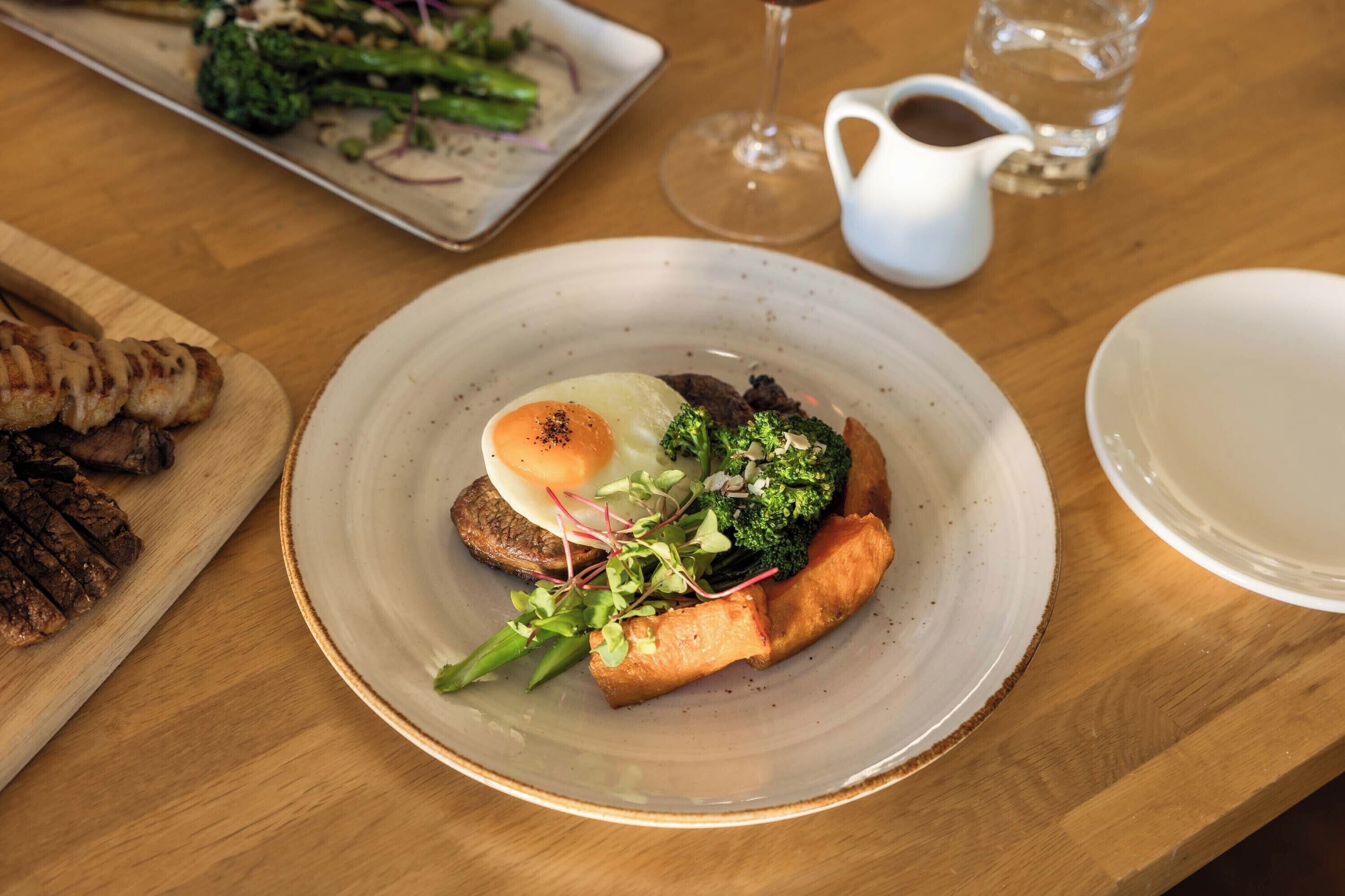 A plate of food with a poached egg, broccoli, and a piece of meat on a wooden table.