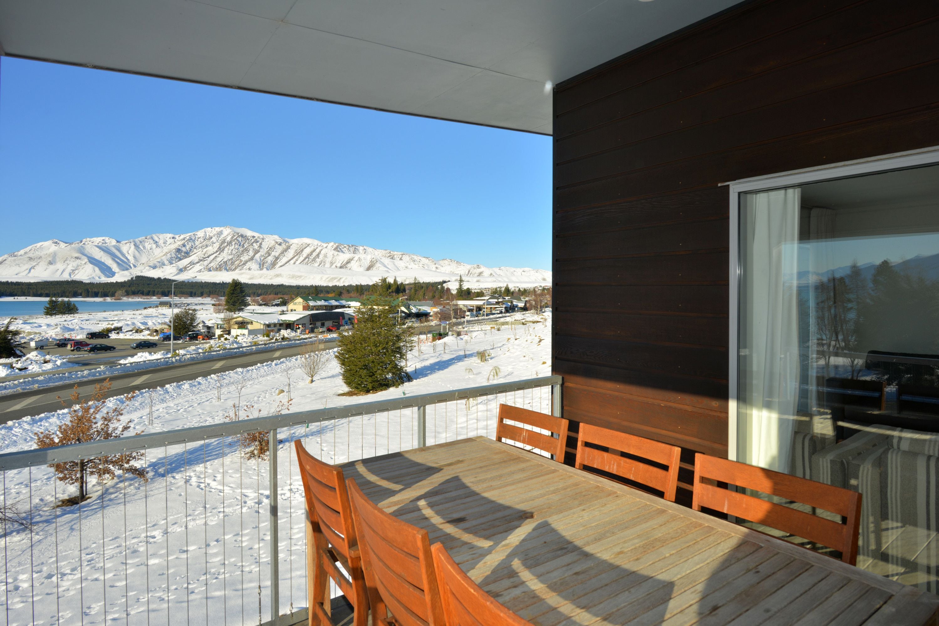 A balcony with a wooden table and chairs, overlooking a snowy landscape with mountains and a clear sky.