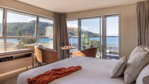 Ett hotellrum med en stor säng, två stolar, ett litet bord och utsikt över en sjö och berg.